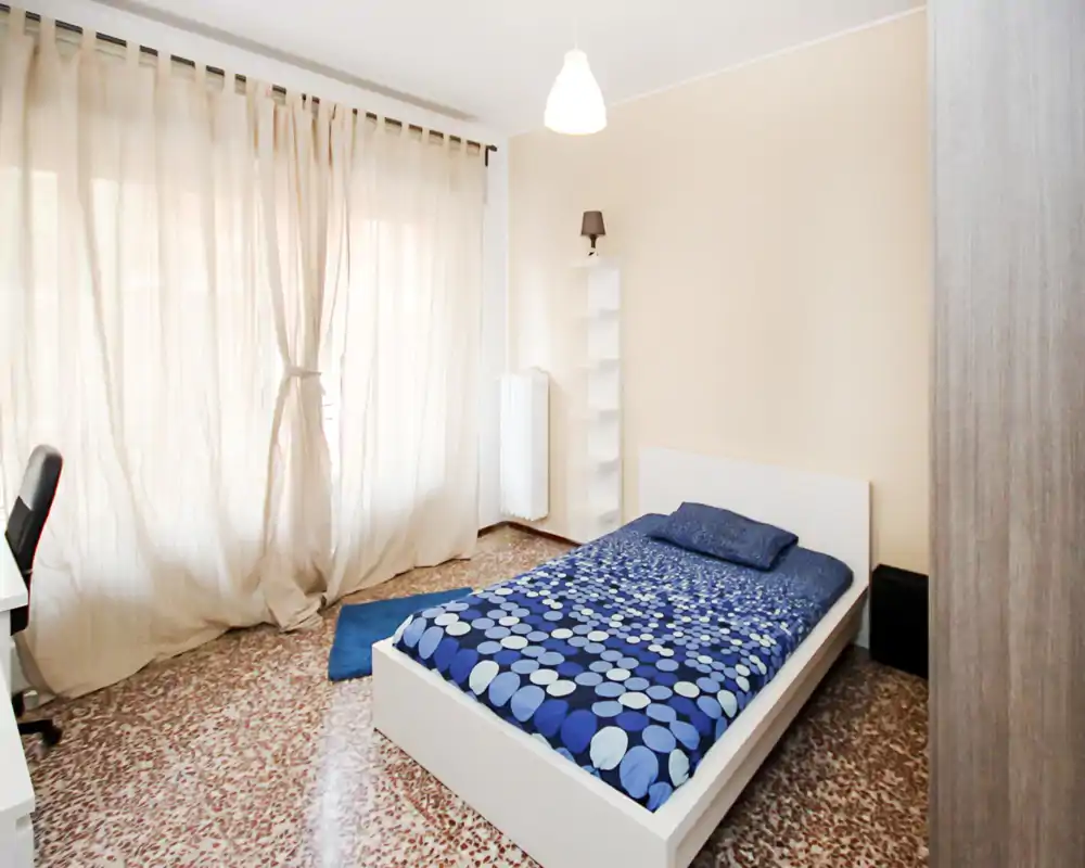 Bright, modern single bedroom with a low white bedframe, patterned blue bedding and large window with sheer curtains letting in soft natural light.