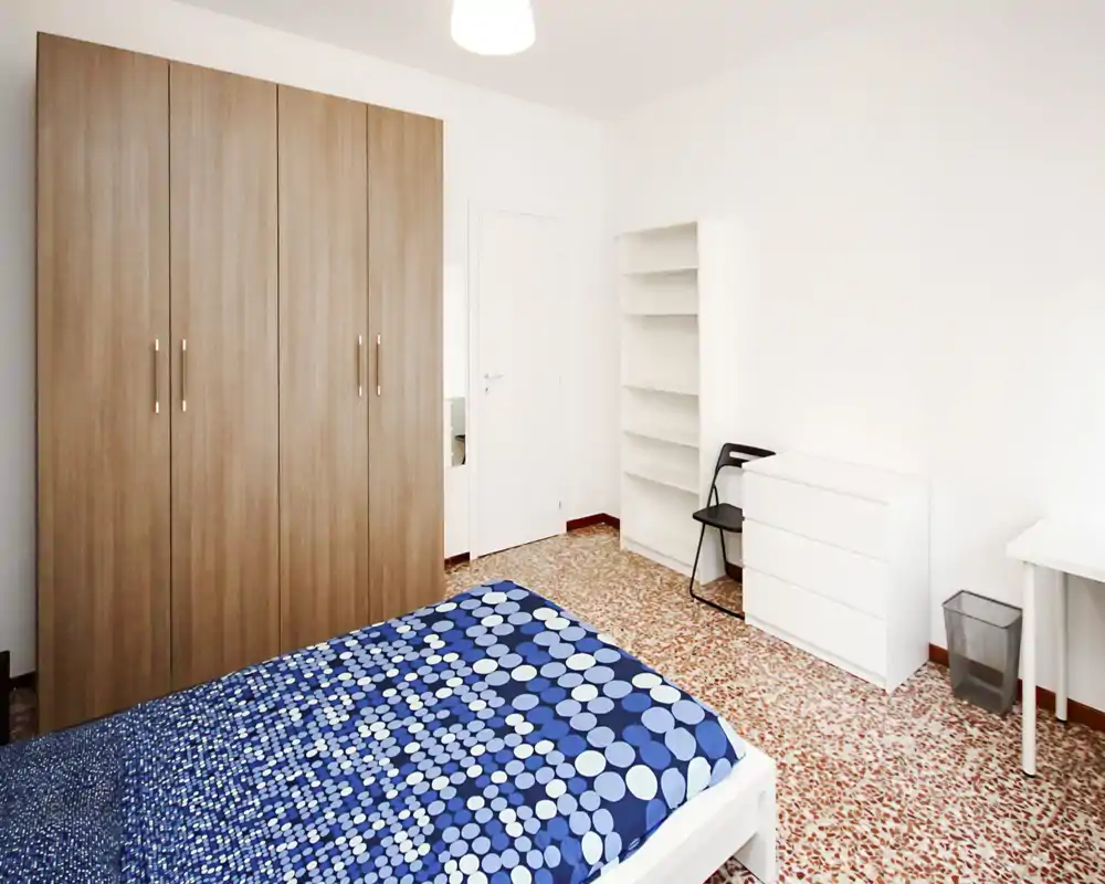 Bright, neatly staged bedroom with a patterned blue duvet, large wardrobe, shelving and simple white furniture — ideal for rental photos.