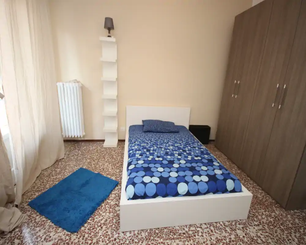 Simple, well-lit single bedroom with a white platform bed, blue patterned bedding, wardrobe and a window providing natural light — neat and ready for guests.