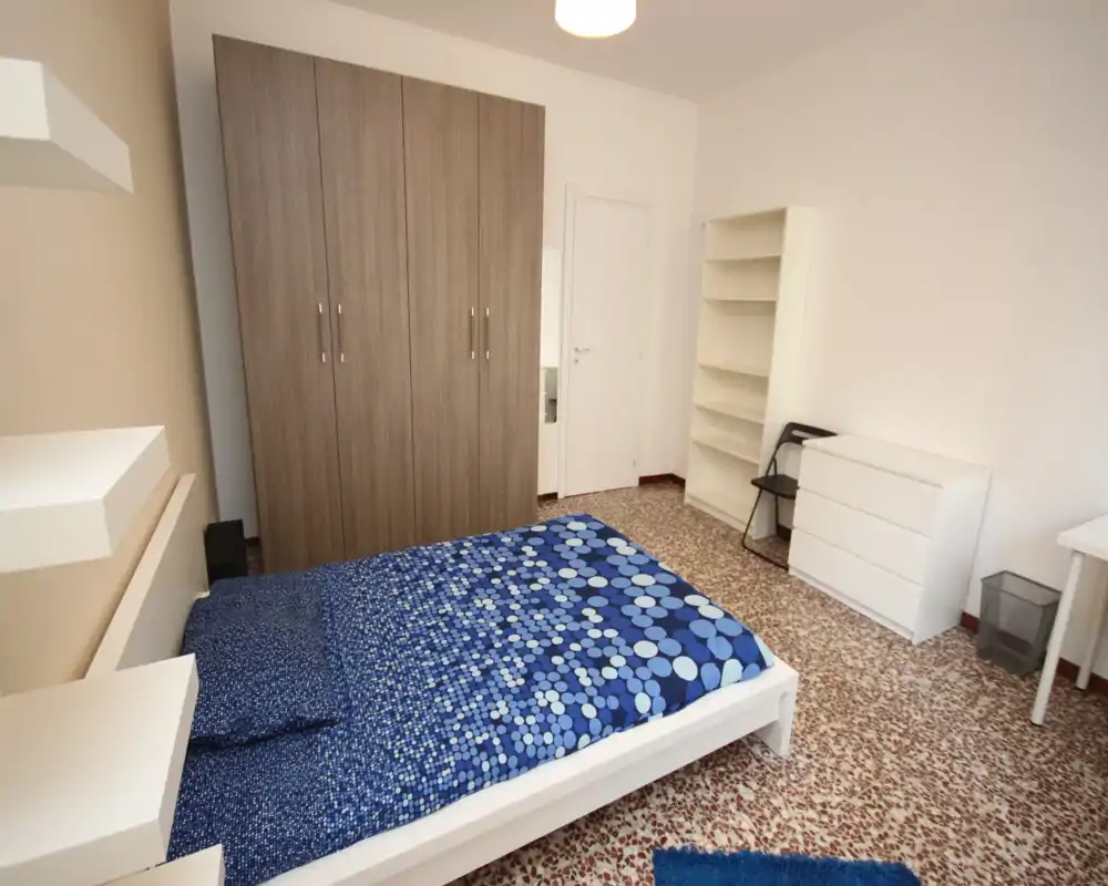 Bright, neatly arranged bedroom with a double bed dressed in a blue patterned duvet, a large wooden wardrobe, open shelving and a white chest of drawers — ideal for rentals looking clean and simple.