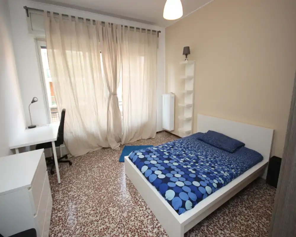 Bright, simply furnished bedroom with a double bed, desk and natural light from a large window with sheer curtains — clean, modern lines and tiled floor.