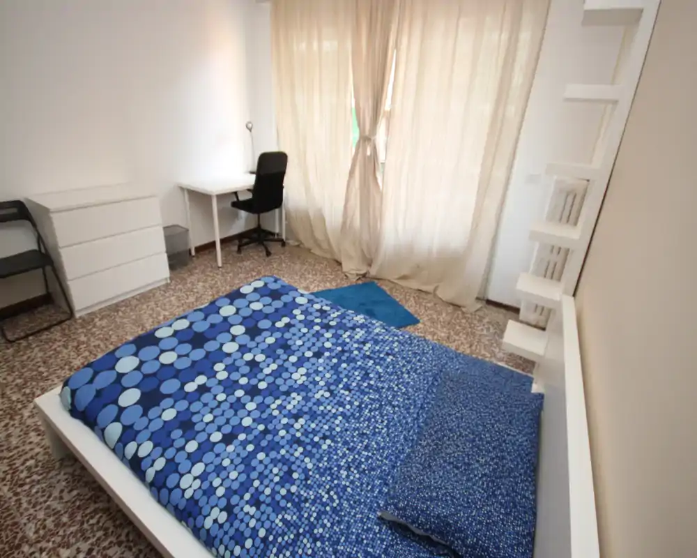 Bright, tidy bedroom with a low white bedframe and blue patterned bedding, a desk and chair by the window, and simple modern storage — ideal for a single occupant or student.