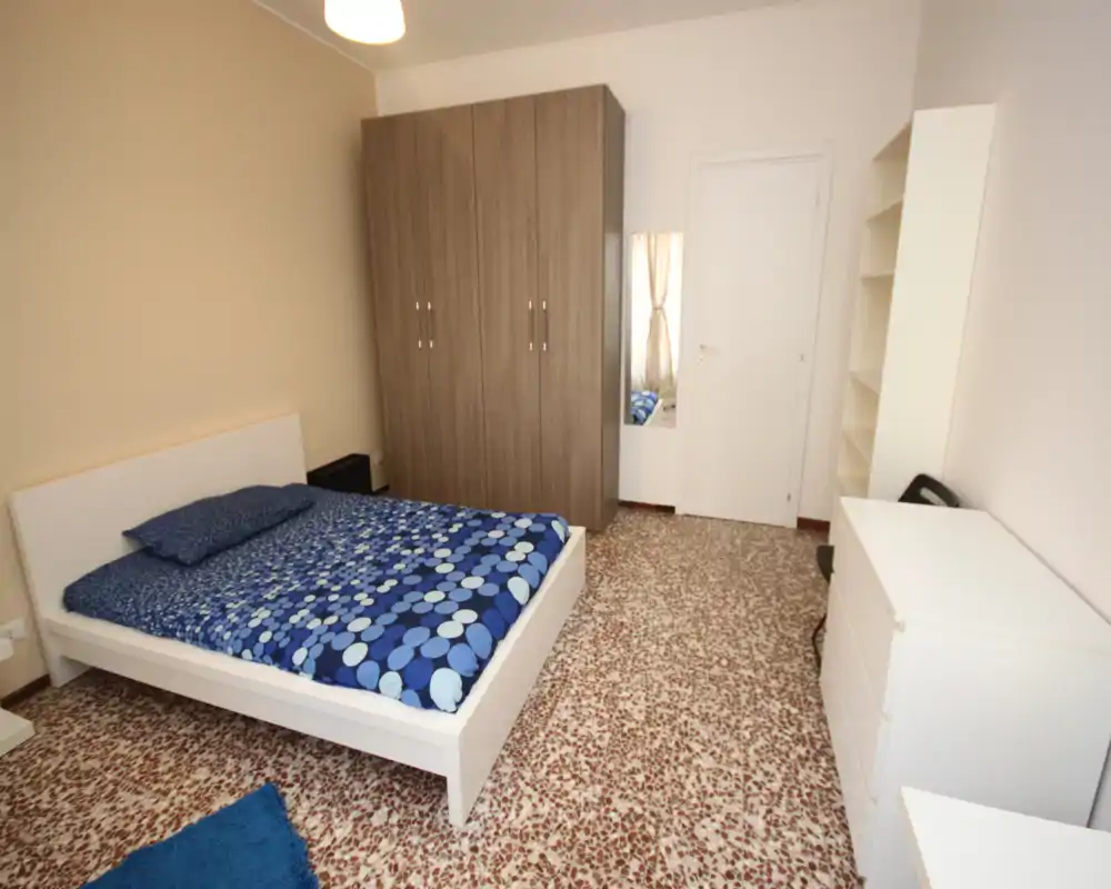 Bright, tidy bedroom with a double bed featuring blue patterned bedding, a large wardrobe, and simple white storage furniture — ideal for a relaxing stay.