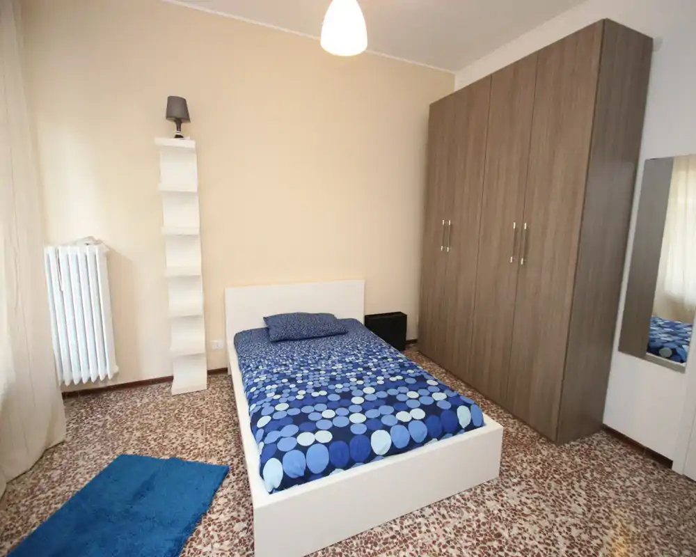 Single bedroom with a modern white bed and blue patterned bedding, large wardrobe, and a simple shelving lamp; bright, tidy and ready for a tenant.