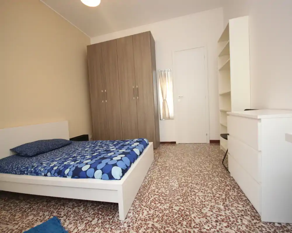 Bright, simply furnished bedroom with a white platform bed, patterned blue bedding, a large wardrobe and shelving—clean and uncluttered, suitable for rental photos.