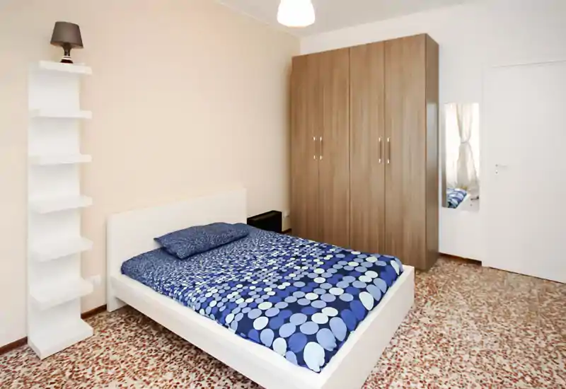 Dormitorio luminoso y ordenado con cama doble con ropa de cama azul estampada, un gran armario y una lámpara de pie — sencillo, moderno y acogedor.