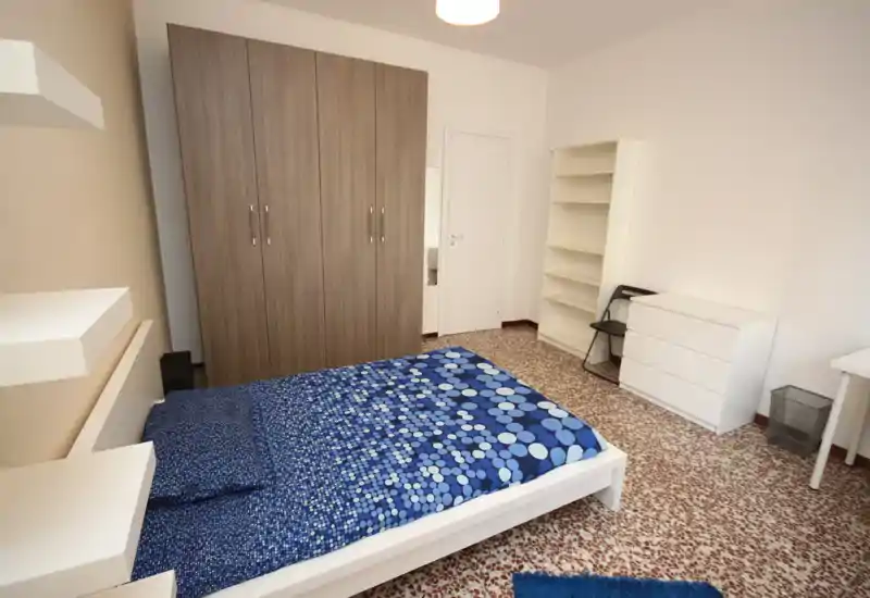 Dormitorio luminoso y ordenado con cama doble con edredón azul estampado, gran armario, estanterías abiertas y cómoda blanca — ideal para anuncios de alquiler limpios y sencillos.