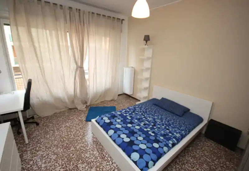 Dormitorio luminoso y ordenado con cama doble con ropa de cama azul estampada, escritorio junto a la ventana y luz natural suave filtrada por las cortinas.