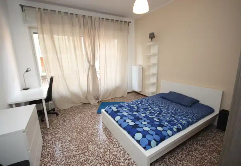 Dormitorio luminoso y de mobiliario sencillo con cama doble, escritorio y mucha luz natural gracias a una gran ventana con cortinas translúcidas — líneas limpias y suelo de baldosa.