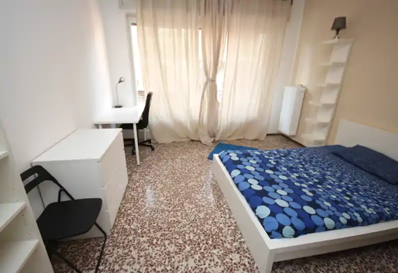 Dormitorio luminoso y de muebles sencillos con cama doble y ropa de cama azul estampada, escritorio junto a la ventana y muebles blancos. La luz natural filtrada por las cortinas aporta una sensación limpia y ventilada, ideal para alquileres temporales.