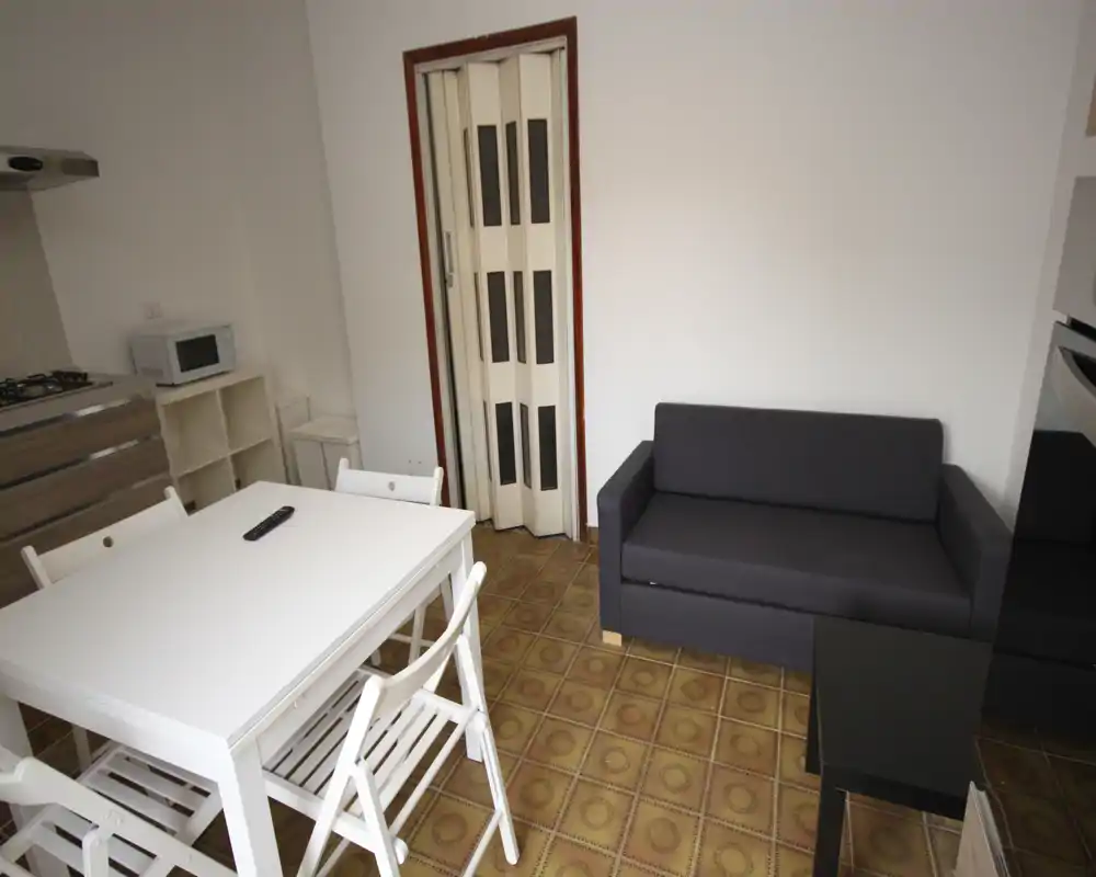 Área de estar/comedor compacta con un sofá pequeño oscuro, mesa blanca con sillas y cocina integrada a la izquierda. Distribución práctica para un estudio, suelo de baldosas y paredes neutras.