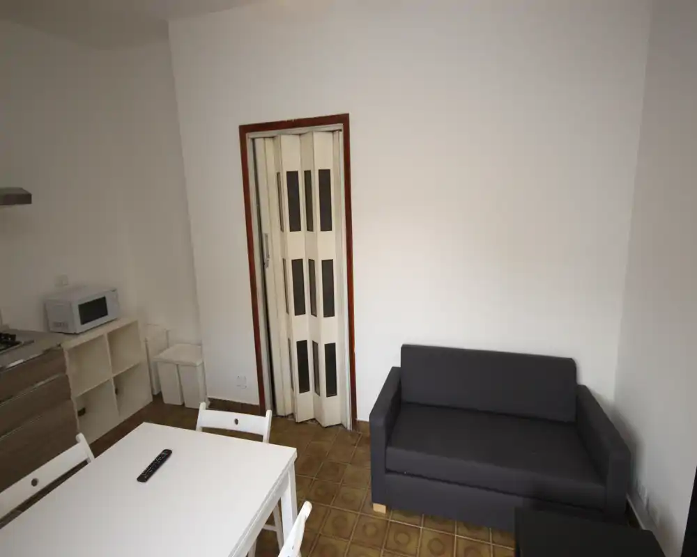 Zona de estar compacta con un pequeño sofá oscuro de dos plazas, una mesa de comedor blanca con sillas y una esquina de cocina. Paredes blancas y suelo de baldosas crean un espacio simple y funcional.