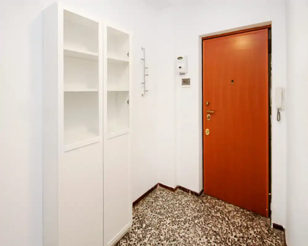 Pequeño recibidor bien iluminado con puerta de madera, portero eléctrico y un armario blanco alto — limpio, minimal y funcional.