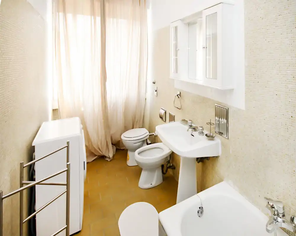 Baño luminoso y limpio con bañera en primer plano, lavabo de pedestal, bidé, inodoro y lavadora junto a una ventana con cortina. Paredes de mosaico neutro y suelo cálido que crean un espacio acogedor y bien iluminado.