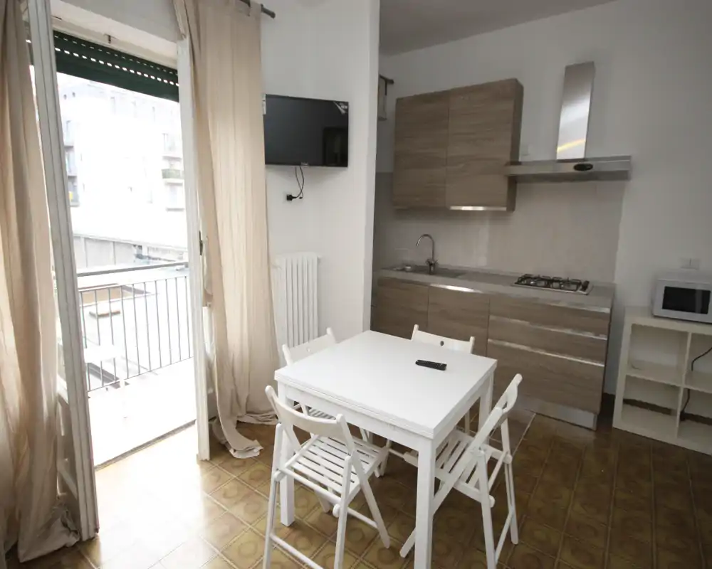 Cocina compacta y luminosa con mesa para cuatro, muebles efecto madera, placa de gas y fregadero. Gran puerta al balcón que aporta luz natural y amplitud.