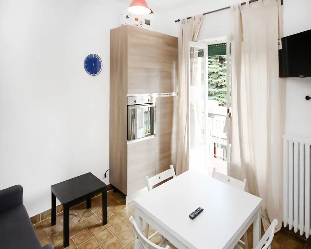 Zona de estar compacta y luminosa con mesa de comedor blanca, sillas y puerta al balcón abierta que aporta luz natural; armario de madera con horno integrado y cortinas claras que crean un ambiente limpio y aireado.