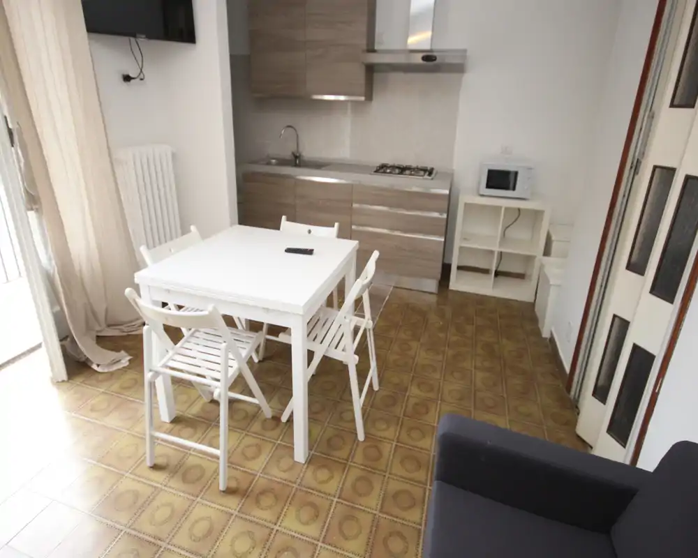 Cocina abierta y compacta con muebles efecto madera, mesa blanca para cuatro y pequeño mueble con microondas. Espacio luminoso y ordenado con luz natural desde el balcón y acabados modernos sencillos.