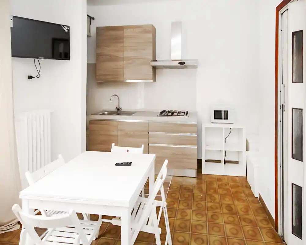 Cocina compacta y luminosa con muebles efecto madera, fregadero de acero inoxidable y placa de gas; una mesa blanca en primer plano crea un espacio funcional y limpio.