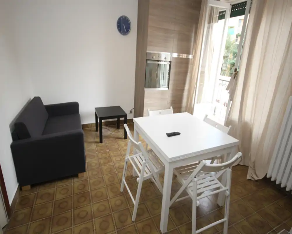 Zona de estar luminosa y ordenada con mesa de comedor pequeña, cuatro sillas y un sofá compacto junto a un balcón que aporta luz natural.