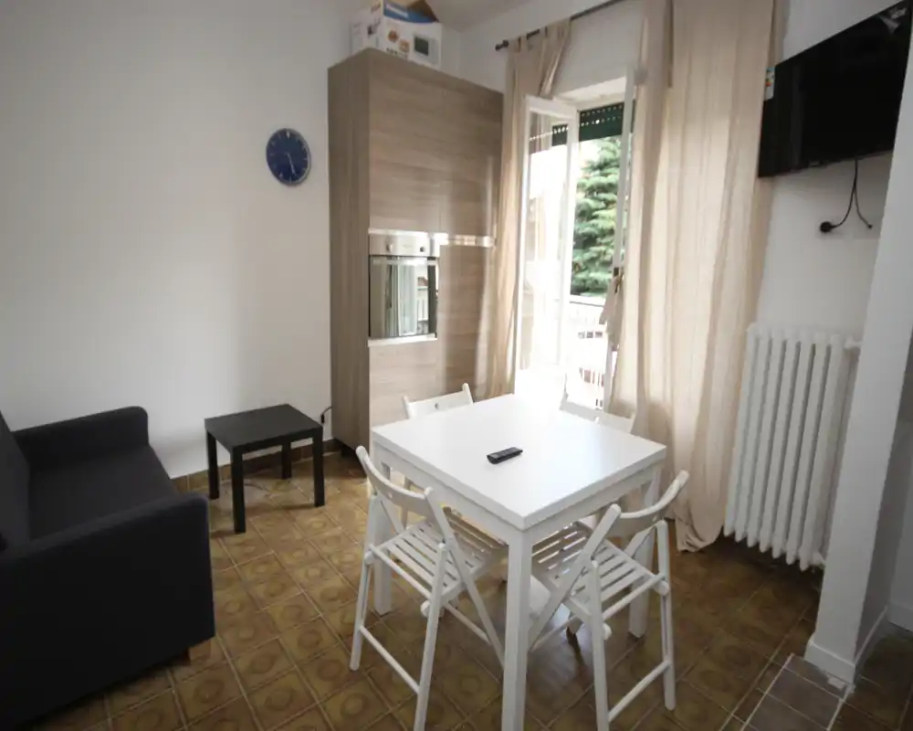 Zona de estar compacta con mesa para cuatro, un sofá y luz natural desde la puerta del balcón. Tonos neutros y mobiliario sencillo crean un espacio práctico y luminoso.