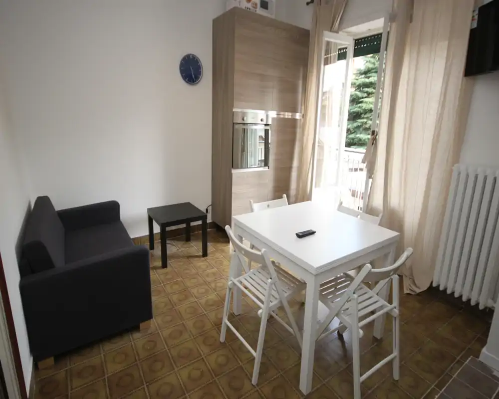 Agradable sala de estar compacta y luminosa con mesa blanca, cuatro sillas, un pequeño sofá y mueble de pared; acceso al balcón con luz natural.