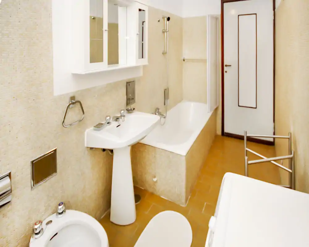 Baño luminoso y cuidado con bañera, lavabo de pedestal, bidé y armario con espejo en la pared; azulejos mosaico beige y suelo de tonos cálidos.