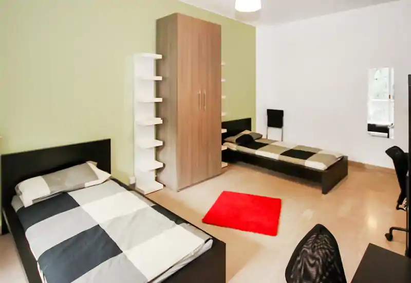 Chambre double moderne et lumineuse avec deux lits simples, une armoire centrale et une touche de couleur grâce au tapis rouge — propre, épurée et bien éclairée.