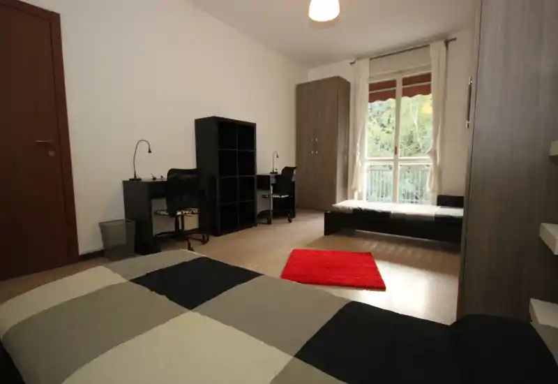 Chambre spacieuse et lumineuse avec deux lits simples, un coin bureau, une armoire et une fenêtre-balcon apportant la lumière naturelle — décoration moderne et tapis rouge qui ajoute de la chaleur.