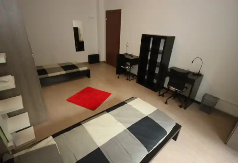 Chambre spacieuse avec deux lits simples, une étagère et deux bureaux ; tons neutres et tapis rouge apportent une touche moderne et sobre.