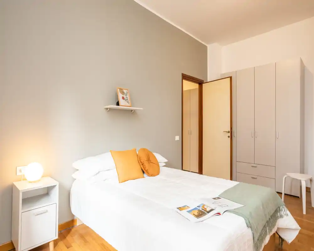 Dormitorio luminoso y bien preparado con cama doble, cojines decorativos cálidos y lámpara de noche; tonos neutros y suelo de madera generan una atmósfera tranquila.