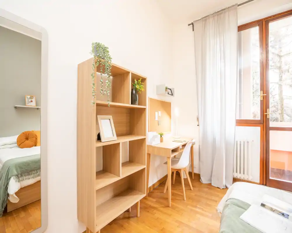 Dormitorio luminoso y acogedor con estantería de madera, escritorio y cama con textiles suaves; gran ventana que aporta mucha luz natural.