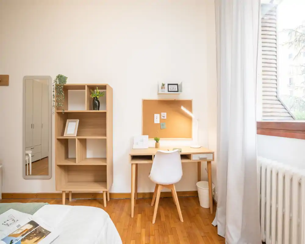 Rincón de dormitorio luminoso y acogedor con escritorio moderno de madera, corcho en la pared y cama parcialmente visible en primer plano. La luz natural de la ventana resalta el suelo de parquet y el mobiliario de estilo escandinavo.