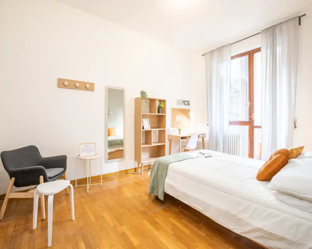Dormitorio luminoso y aireado con cama doble con ropa blanca, suelo de madera cálido, butaca de lectura y un pequeño escritorio junto a la ventana — ideal para estancias cómodas.