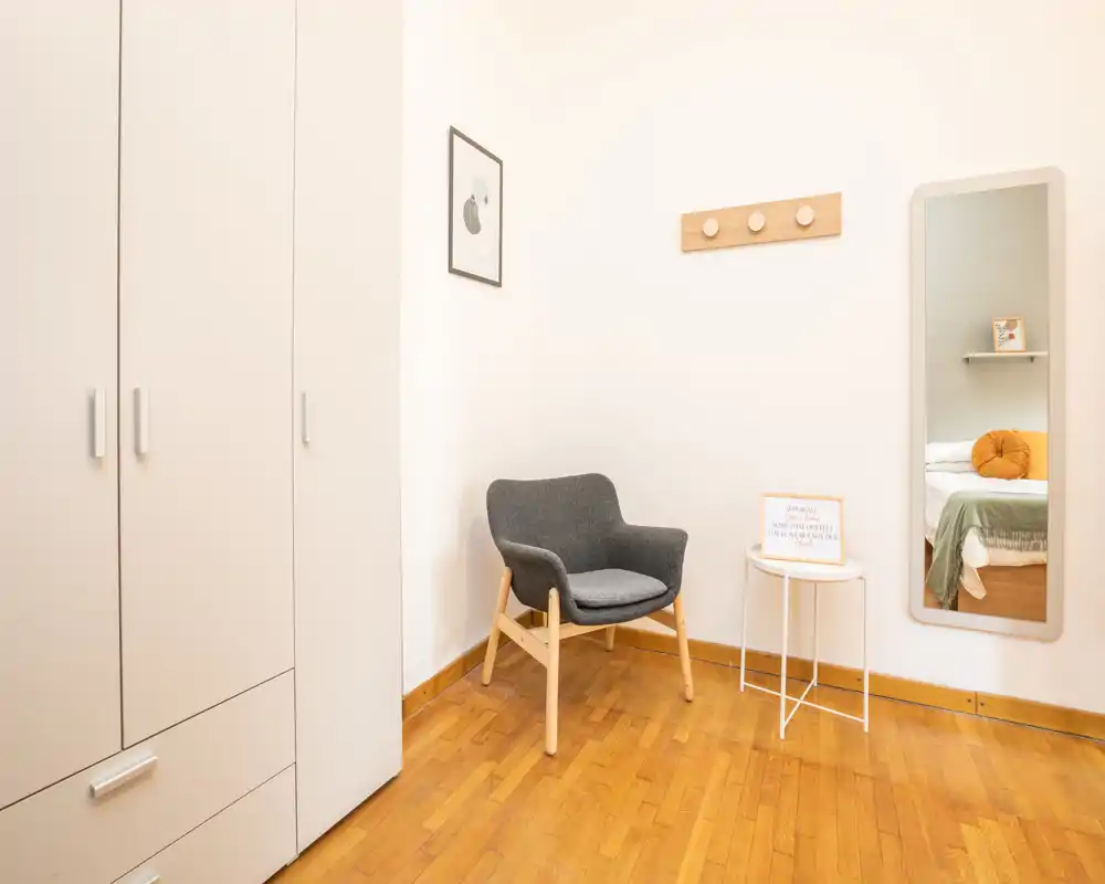Rincón de dormitorio luminoso y minimalista con armario, butaca y espejo de cuerpo entero que refleja parte de la cama; suelos de madera y decoración neutra que transmiten calma.