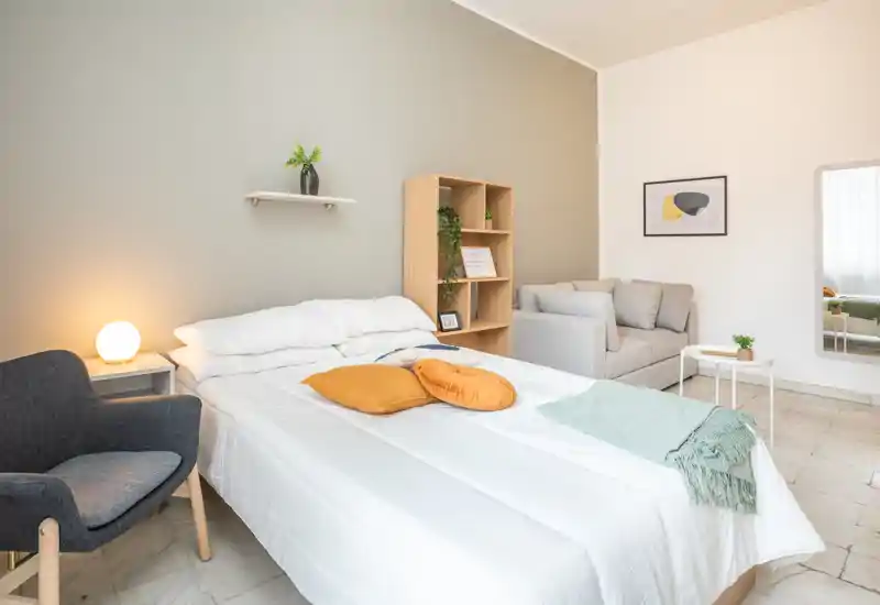 Dormitorio moderno y luminoso con cama doble bien hecha, cojines acogedores y una manta; incluye un sillón, un pequeño sofá y estantería, ideal para anuncios de alquiler.