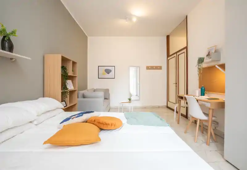 Dormitorio luminoso y bien decorado con una cama grande en primer plano, cojines cálidos, un sofá pequeño y una zona de trabajo compacta: ideal para una persona o pareja que busca una habitación acogedora y funcional.