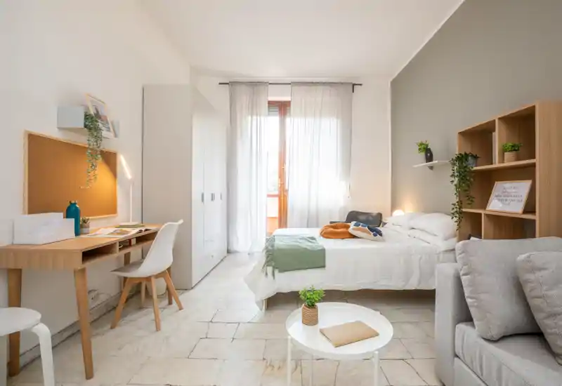 Dormitorio luminoso y bien decorado con cama doble, zona de escritorio y acceso al balcón. Ropa de cama suave, cojines y plantas generan un ambiente acogedor perfecto para anuncios.