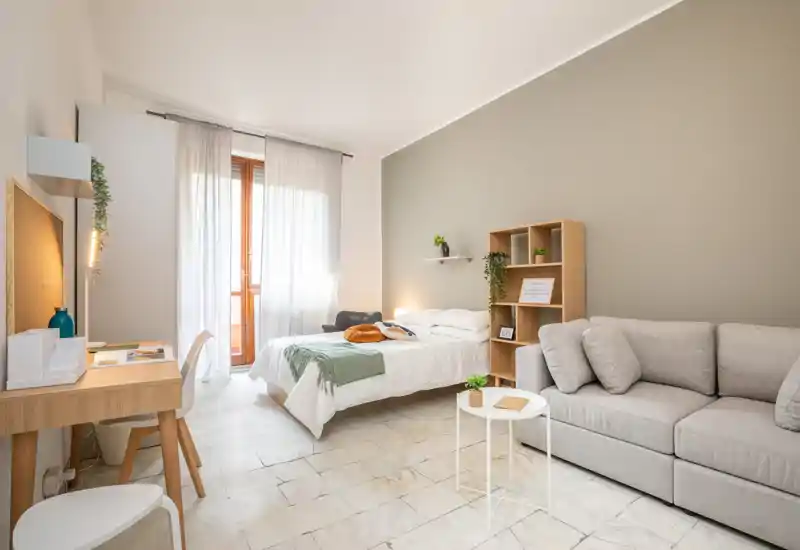 Estudio luminoso y bien decorado con una cama doble cómoda, textiles suaves y un área de estar. La luz natural desde la puerta al balcón y las estanterías de estilo sencillo generan un ambiente cálido y acogedor.