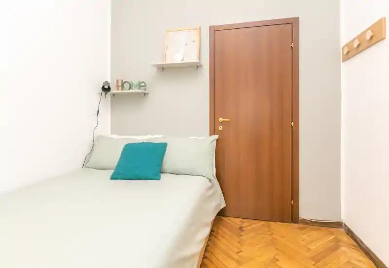 Small, neatly made bedroom with a double bed, pastel bedding and a teal accent cushion, warm wooden door and parquet floor creating a cozy, minimal atmosphere.