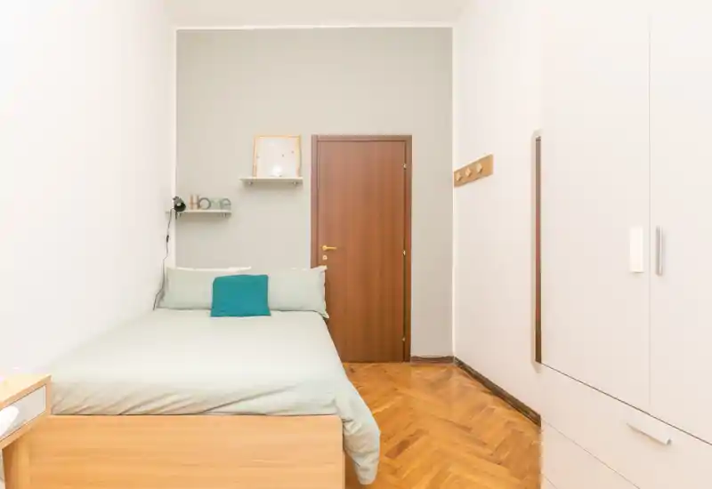 Bright, minimal single bedroom with a neatly made bed, wooden parquet flooring and a built-in wardrobe. The room has soft green bedding, a small shelf with decor and a warm wooden door, ideal for a cozy rental listing.