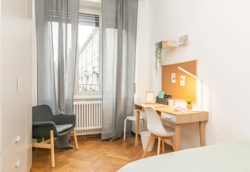 Bright, cozy bedroom corner with a wooden desk and a comfortable chair by a large window; soft natural light and minimalist decor make it ideal for a pleasant stay.