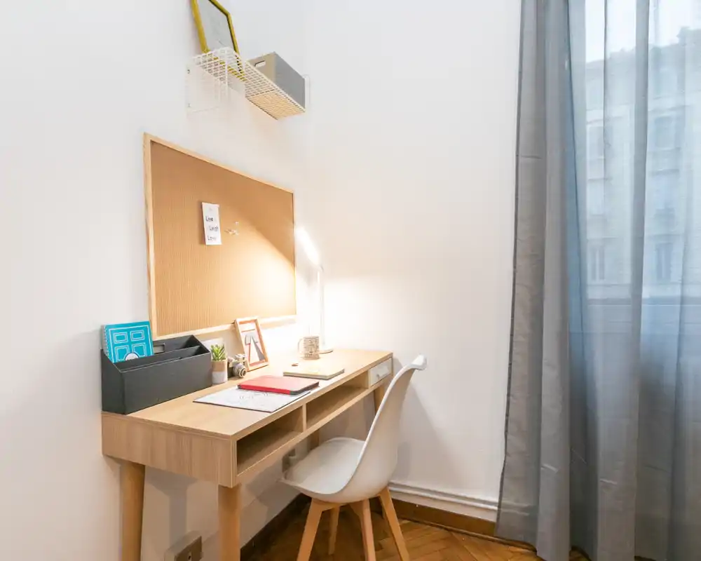 Espacio de trabajo luminoso y minimalista con escritorio de madera, panel de corcho, lámpara de escritorio y silla blanca junto a una ventana con cortina translúcida — ideal para trabajar o estudiar.