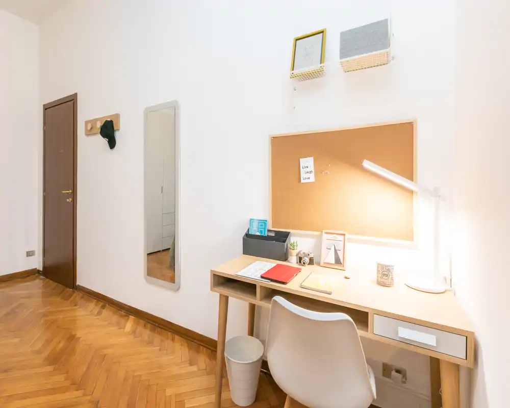 Rincón de trabajo luminoso en el salón: escritorio de madera con lámpara, corcho y silla sobre paredes blancas y parquet en espiga.