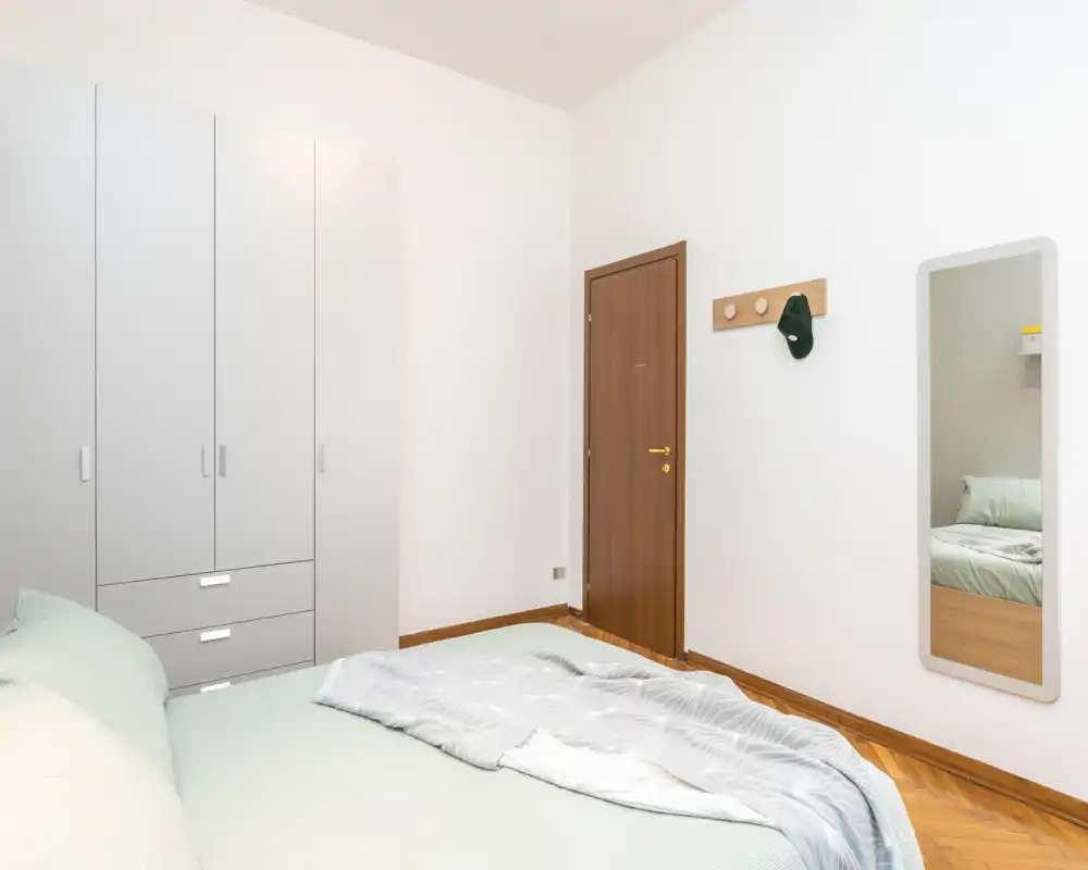 Dormitorio luminoso y minimalista con una cama doble bien hecha, ropa de cama clara y un gran armario blanco. Decoración sencilla con espejo en la pared y puerta de madera, ambiente limpio y aireado perfecto para anuncios.