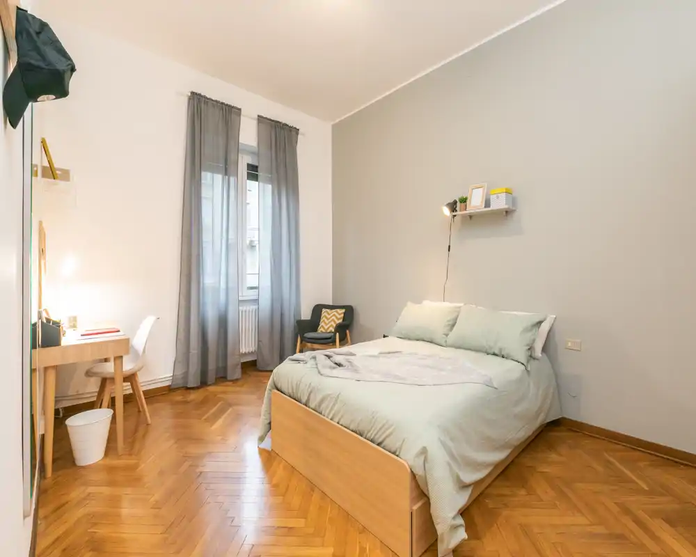 Dormitorio moderno y luminoso con cama doble, ropa de cama verde suave, suelo de parquet y un pequeño escritorio junto a la ventana — bien iluminado y acogedor.