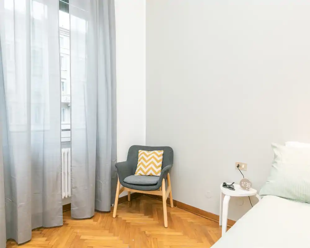 Rincón de dormitorio luminoso y minimalista con un sillón acogedor, mesita de noche y cama parcialmente visible con ropa de cama suave — ideal para una estancia tranquila.