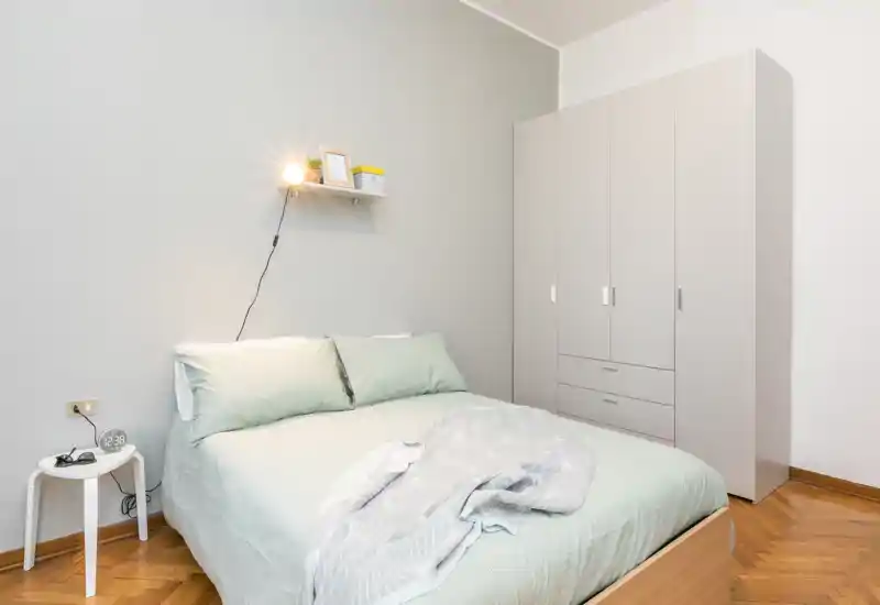 Dormitorio luminoso y sencillo con cama doble claramente visible, mesita y armario independiente. Tonos neutros y suelo de madera cálida crean un ambiente tranquilo y acogedor, perfecto para anuncios de alquiler.