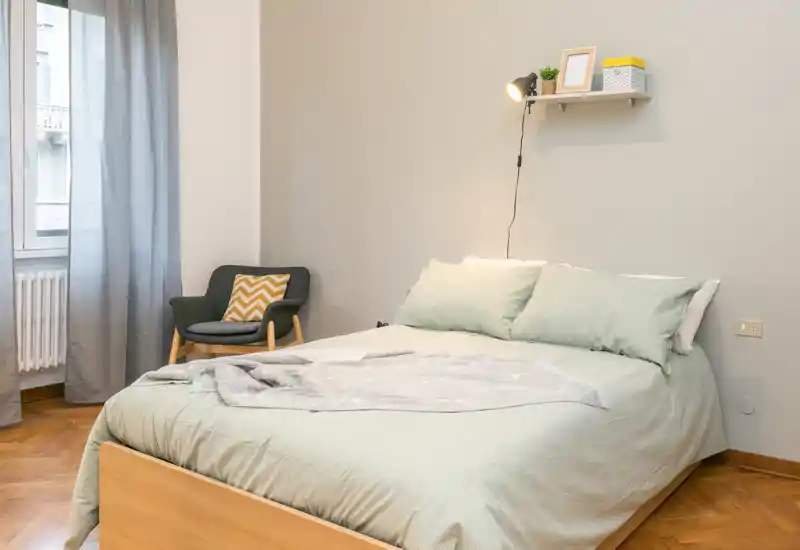 Dormitorio luminoso y moderno con una cómoda cama doble con ropa de cama verde claro, una lámpara de pared y un pequeño sillón junto a la ventana — ideal para estancias relajantes.