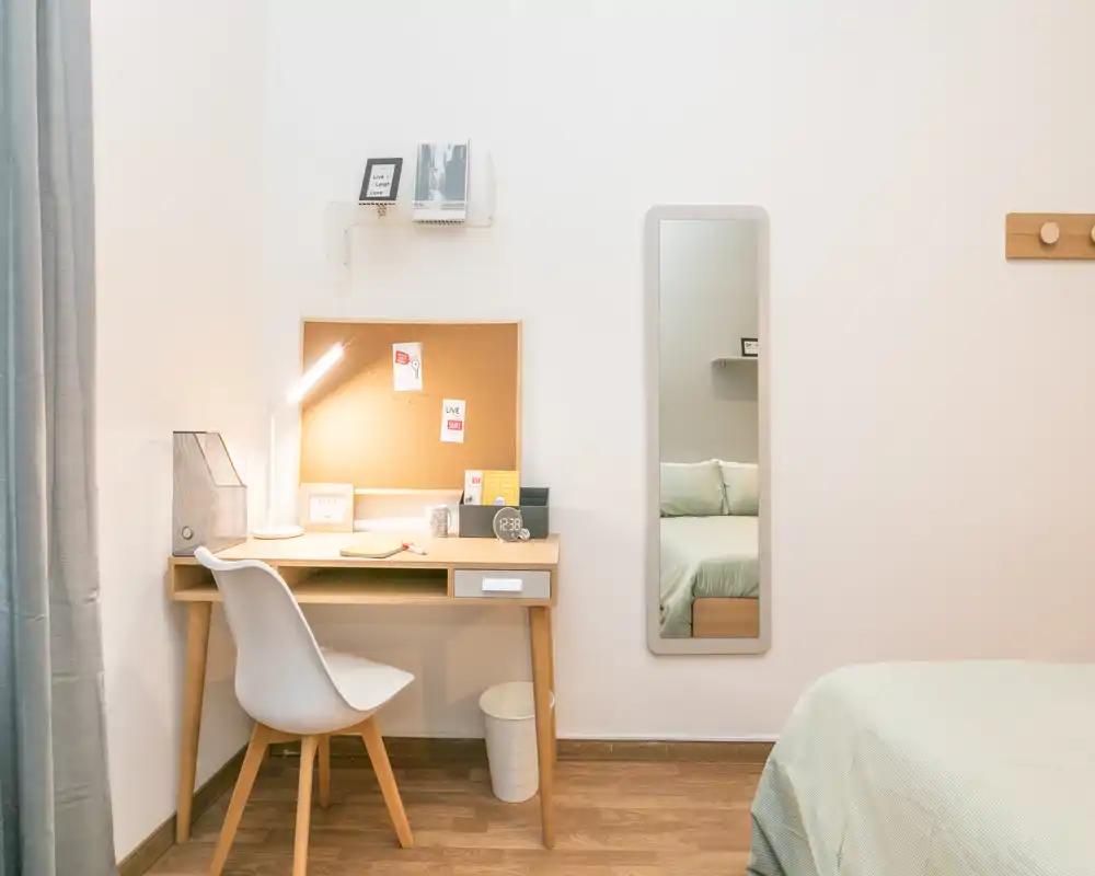 Rincón de dormitorio luminoso y minimalista con escritorio de madera, lámpara de lectura acogedora y cama parcialmente visible con ropa de cama suave — ideal para una persona o estudiante.
