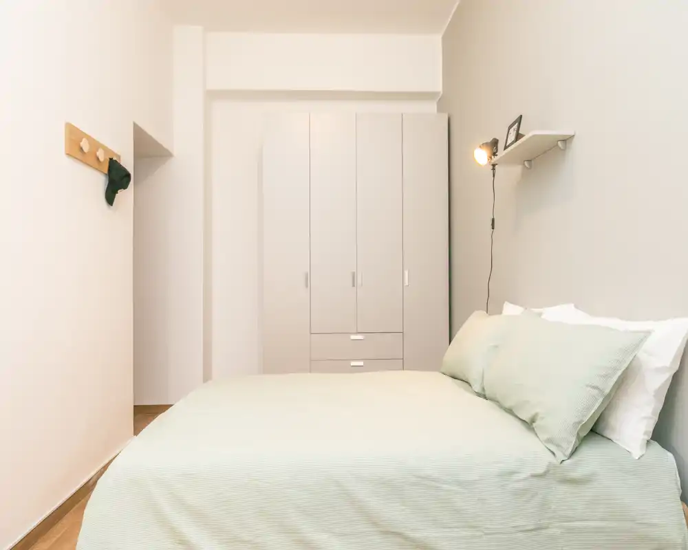 Dormitorio luminoso y minimalista con cama doble de ropa de cama verde pálido, armario independiente y lámpara de pared: espacio tranquilo ideal para un anuncio.